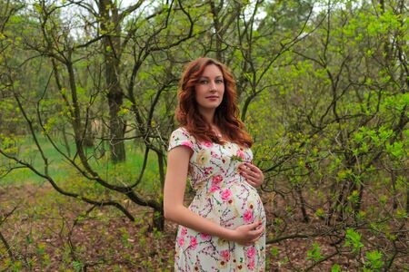 Portrait of beautiful pregnance woman near the green tree. Red hair, lips. Mother and babyの写真素材