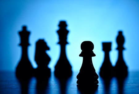 Creative image depicting playing chess with a low angle view of chess pieces on a reflective surface against a graduated blue background with backlighting and focus to a single pawnの写真素材