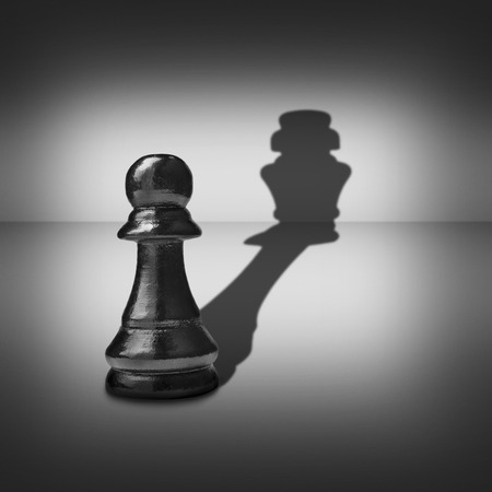 Conceptual image of dual identity with a pawn chess piece casting a shadow belonging to a king across a central highlight surrounded by a vignette in a black and white imageの写真素材