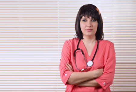 Beautiful confident young female doctor standing with a stethoscope around her neck looking intently at the camera with folded armsの写真素材