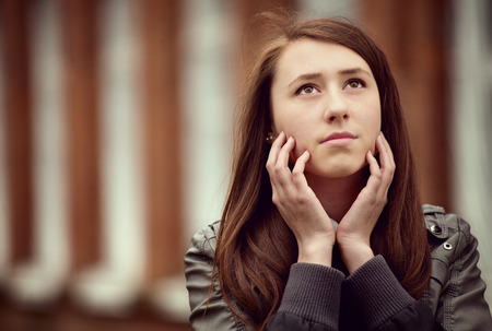 Thoughtful young woman sitting daydreaming with her hands raised to her cheeks staring meditatively...