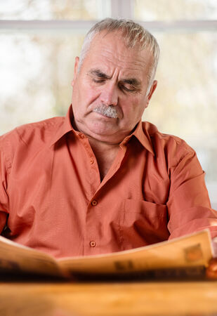 Portrait of a Caucasian senior man wearing mustache and a brick-red shirt while sitting at the table reading the daily newspaper with a serios and worried facial expressionの写真素材