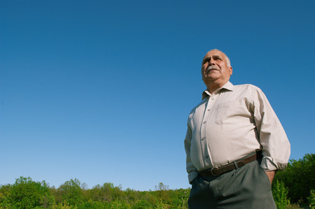 Low angle view of an overweight senior man standing with his hands in his pockets against a blue sunny sky in the countryside accentuating his bellyの写真素材