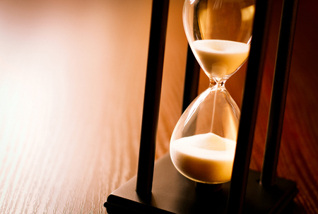 Hourglass with the sand running through in a wood frame on a wooden surface with shine and copyspace