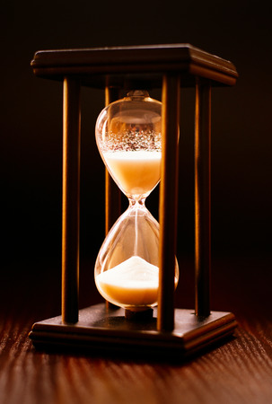 Illuminated hourglass in a wooden frame with sand pouring through between the glass bulbs measuring the passing time against a dark backgroundの写真素材