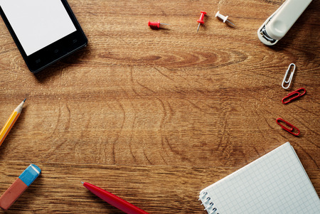 Frame of stationery and office supplies with a tablet, stapler, thumbtacks, paperclip, notebook, pen, pencil and eraser arranged on a wooden textured background with central copyspaceの写真素材
