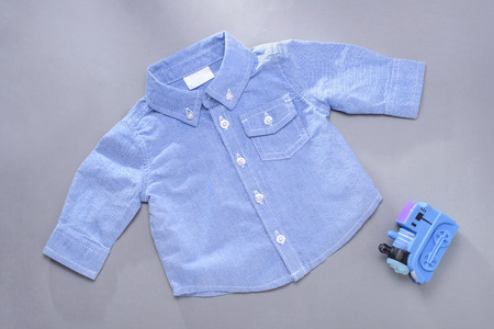Single blue infant buttoned long sleeve shirt with front pocket next to plastic toy train locomotive on gray backgroundの写真素材
