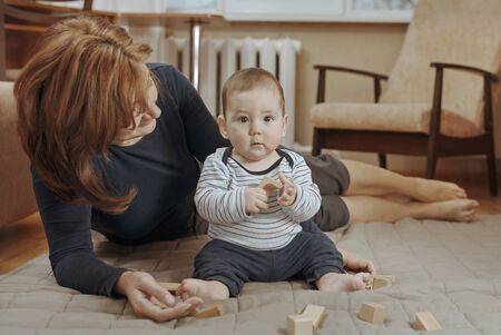 Young mother watching over her cute little baby boy as he plays on the carpet in the living room with wooden blocks in a close up low angle portraitの写真素材