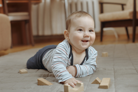 Cute happy little baby boy playing on the carpet at home lying on his stomach looking up to the side...