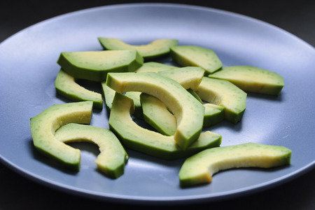 A close up of multiple green avocado slices on a blue plate with a dark background.の写真素材
