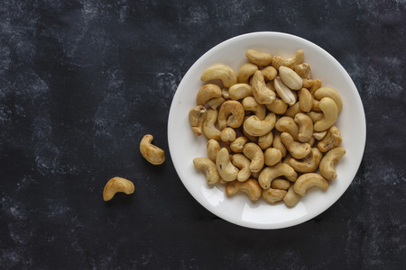Roasted salted cashew nuts on a white plate viewed from above on a black background with copy spaceの写真素材