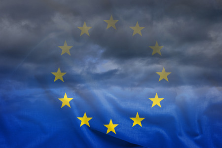 Dark clouds in the sky viewed through half-transparent European Union flag blowing in the wind. Full...