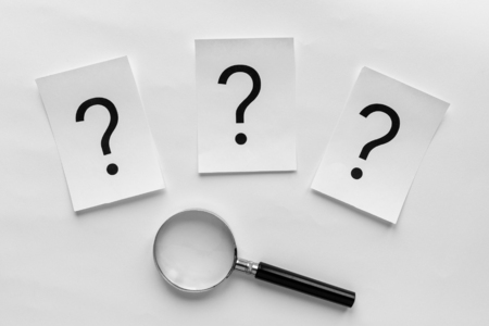 Three question marks printed on white cards in a line above a magnifying glass on a white background...