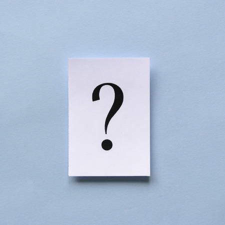Single black printed question mark on a white card or sign with three dimensional shadow over a blue...