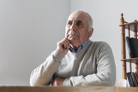 Elderly man with mustache sitting thinking with his hand to his chin staring ahead with a pensive expressionの写真素材