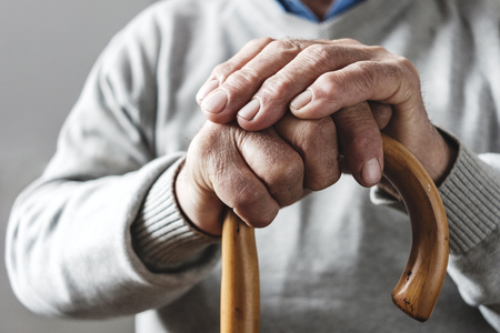 Close up details of the folded hands of an elderly man resting on a walking cane in a mobility and...