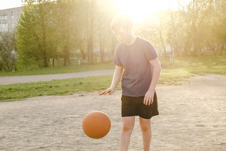 The boy plays a basketball in the open air in the rays of the evening sun.