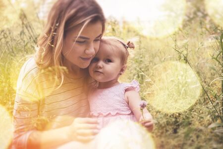 Loving mother playing outdoors with her cute little baby daughter amongst the spring grass as the small girl holds up a purple flower with a smileの写真素材