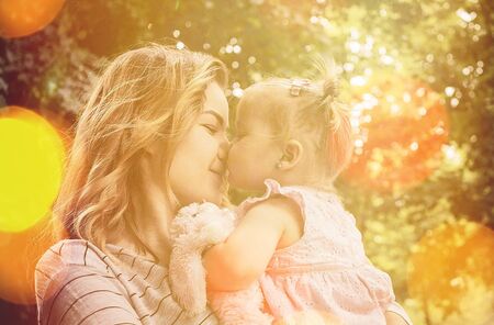 Happy young mother kissing her cute little baby daughter with blond pigtails outdoors in a wooded park amongst treesの写真素材