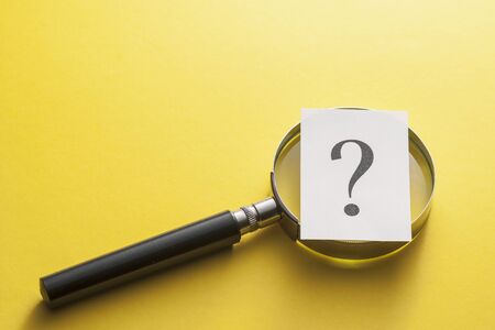 Magnifying glass covered by a question mark printed on card lying diagonally on yellow with copy...