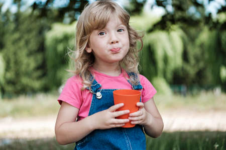 Cute little blond girl pursing her lips as she drinks juice from a colorful red mug outdoors in the garden in summer in close upの写真素材