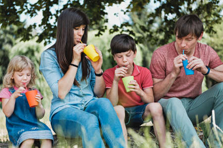 Young family with children relaxing outdoors drinking juice from colorful mugs in the shade of a...