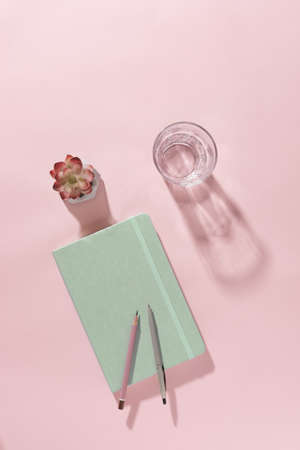 Pastel pink office background with closed green notebook, glass of fresh water with shadow, flower and pen in a business, creativity or personal diary conceptの写真素材