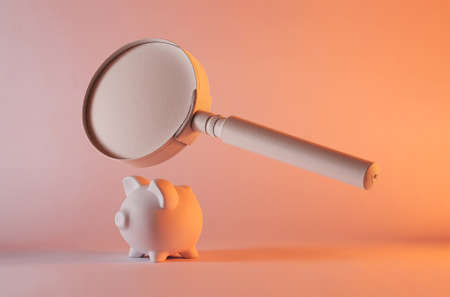 Close-up of a matte old magnifying glass floating above a small piggy bank against studio pink background as symbol for financial investigation and research about saving moneyの写真素材