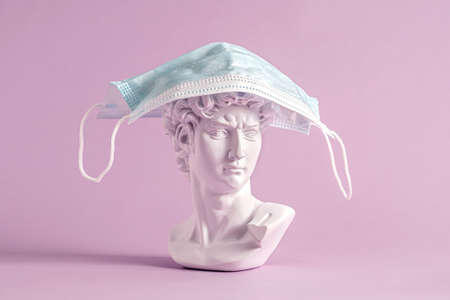White Grecian or Roman bust with face mask draped on top covering the head over a pink background in a conceptual Covid-19 pandemic imageの写真素材