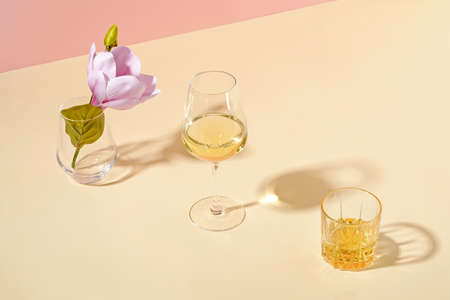 Single fresh summer bloom in vase with glass of white wine casting long shadows over a yellow...