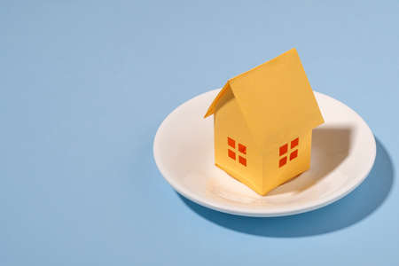 Close-up of a small yellow house made of paper on a plate against blue background for copy space as symbol for residence and real estateの写真素材