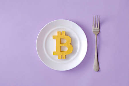 The symbol of the modern digital cryptocurrency Bitcoin in a plate and a fork on the table as food....