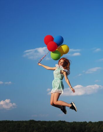 Young redhead girl with colorful balloons jumping at the blue skyの写真素材