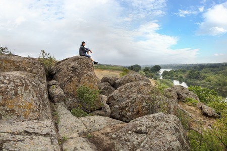 The teenager sits on top of a large stone boulder on the bank of the South Bug River and looks at the river below. The river Southern Bug in the summer - river flow, rocky shores, bright green vegetation and a cloudy blue skyの写真素材