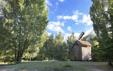 An old wooden windmill on a green glade in a summer young forest, against a blue sky with light white cloudsの写真素材