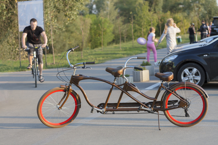 A chocolate-colored tandem bicycle with scarlet wheels is parked in a car park near the bike path in the evening sun.の写真素材
