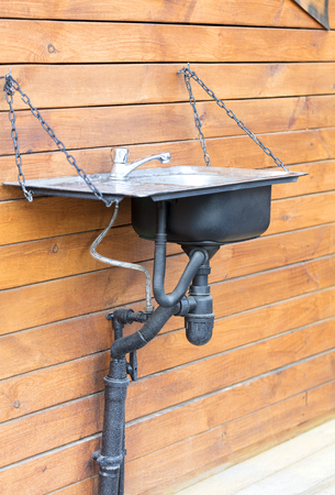 A modern washbasin sink with a stainless steel faucet installed outside on a chain on the wall of a wooden house, in a rustic farmhouseの写真素材