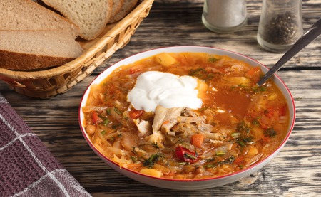 Ukrainian borscht and a spoon of sour cream in a white porcelain plate, a basket with sliced bread and a wafer napkin on an old wooden tableの写真素材