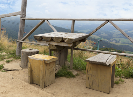 Carpathians. An old wooden weather table and three rough improvised chairs for a respite on top of the mountain. Ski lift in the background of a mountain landscape.の写真素材