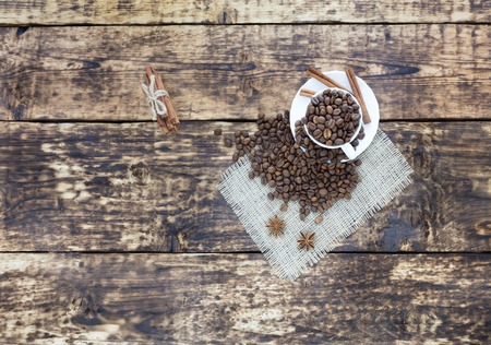 On the old wooden table is a white cup with grains of coffee. Grains of coffee are scattered on a linen napkin. The cinnamon is tied with a rope. Anise complements the aroma of coffee.の写真素材