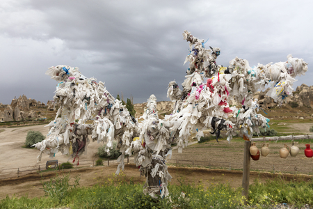 The tree of desires is decorated with white ribbons in Goreme, Central Turkey against the background of the leaden-gray sky and mountain landscapes of Cappadociaの写真素材