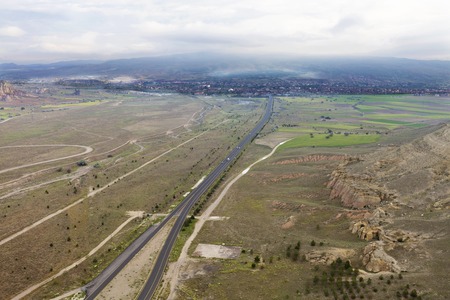 An asphalt road leads to the village through a brown valley and fields between the mountains from a...