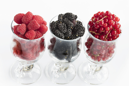 Raspberry, a big black blackberry and red currant are poured into transparent glass cups for latte and stand in a row on a light background, the image in a high keyの写真素材