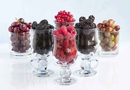 Raspberry, gooseberry, a big black blackberry, red currant and mulberry are poured into transparent glass cups for lattes and stand in a row on a light background, the image in a high keyの写真素材