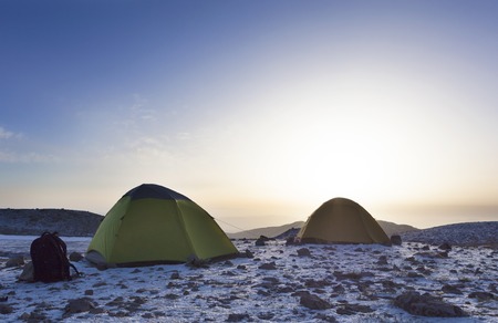 Backpacks and tents of tourists are located on top of the mountain against a clear blue sky and the rising morning sunの写真素材