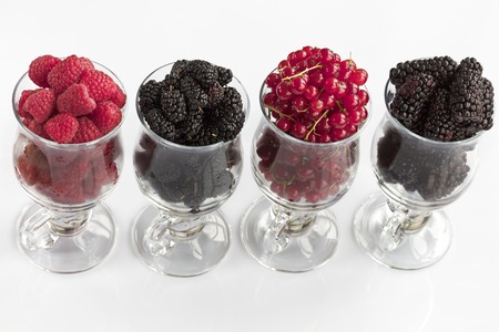 Raspberry, a big black blackberry, red currant and mulberry are poured into transparent glass cups for lattes and stand in a row on a light background, the image in a high keyの写真素材