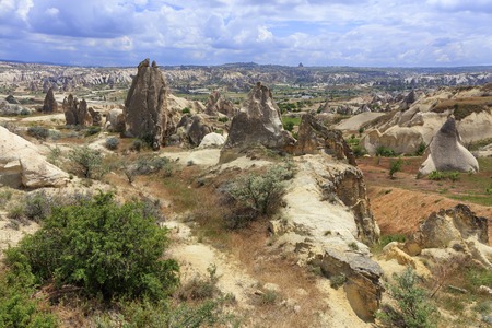 A wild rocky landscape among conic rocks, ancient caves, burnt grass and thorny shrubs runs a winding path against the backdrop of the mountainous landscape of the valley in Cappadocia.の写真素材