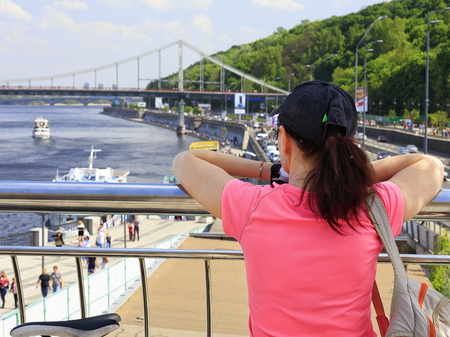A young woman looks at the embankment of the Dnieper River in Kiev, Ukraine. A lady traveler in a bright pink T-shirt stands on the observation deck and looks towards the bridge.の写真素材