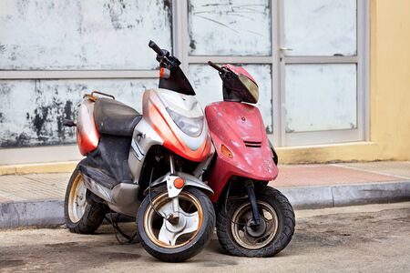 Two old worn scooters, red and silver, stand alone on the side of the road against the background of...