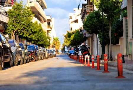 Red reflective road columns separate the pedestrian area of the road from the carriageway amid parked cars, a deserted street and bright morning sunlight in blur.の写真素材